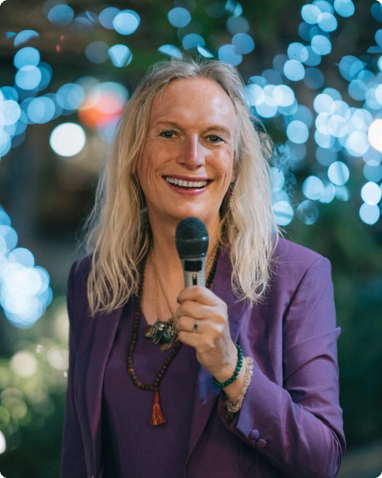 Woman Speaking At Event With Microphone, Smiling, In Front Of Blurred City Lights.